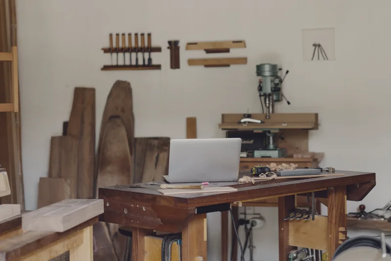 A woodworking shop: an open laptop on the workbench, with boards and tools along the wall.